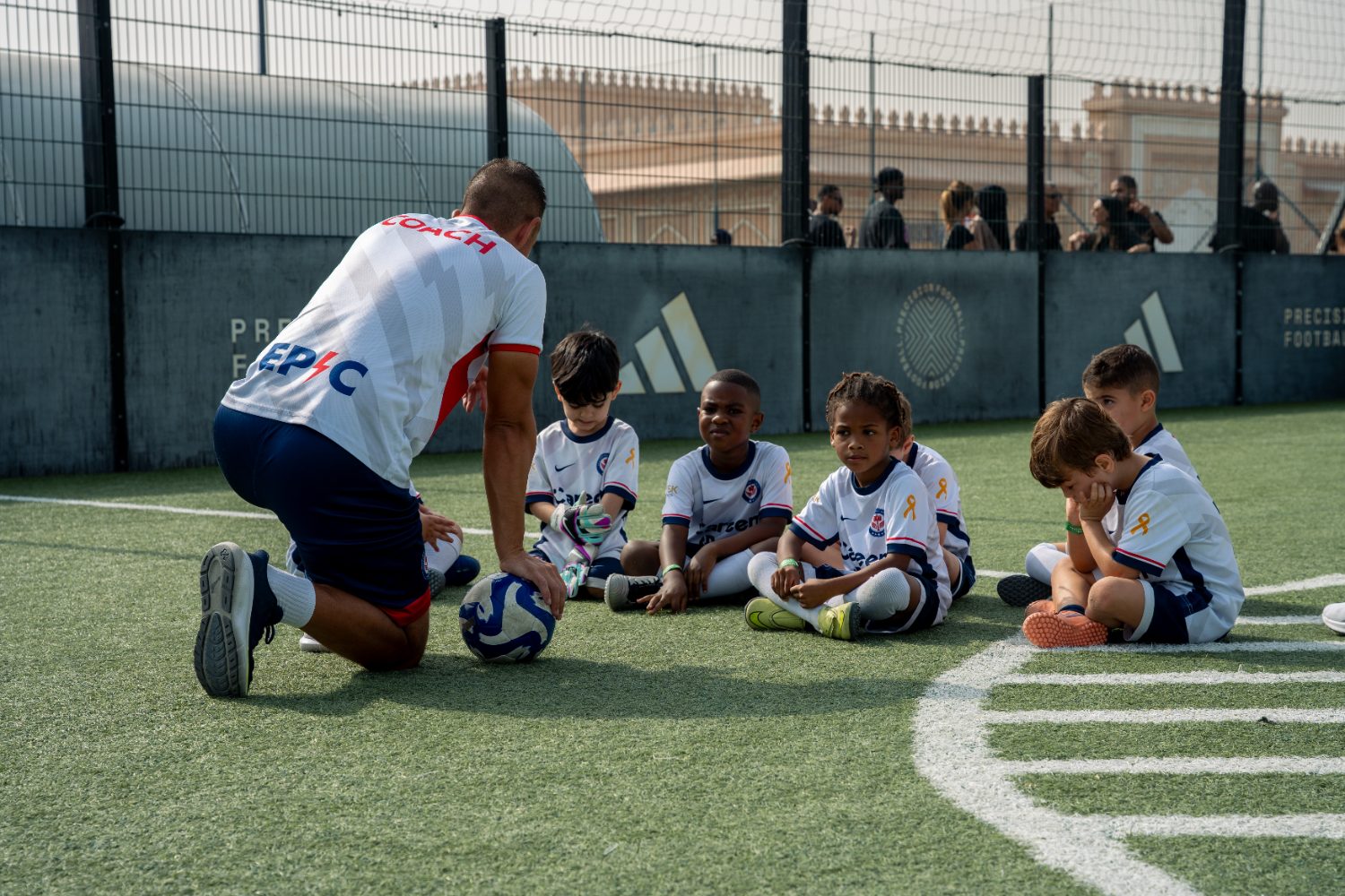 EPIC coach kneeling with a group of young children aged 4 to 6 sitting on the pitch in Dubai